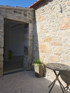 Un mur de pierre avec une porte qui mène à une pièce. On y trouve une petite table avec un pied en métal et, à côté, une plante en pot.