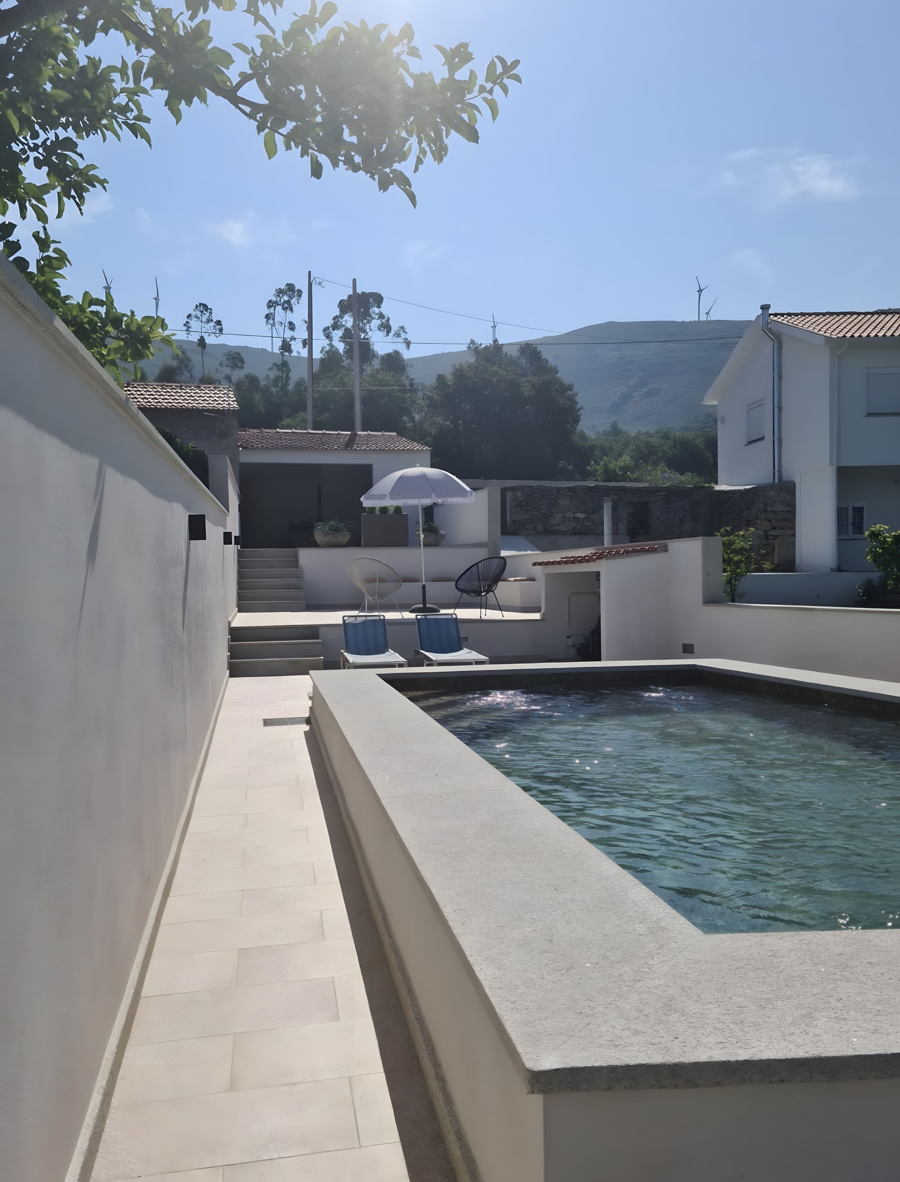 Une piscine bordée de pierres, deux chaises longues et un escalier menant à un espace abrité sous un parasol blanc.