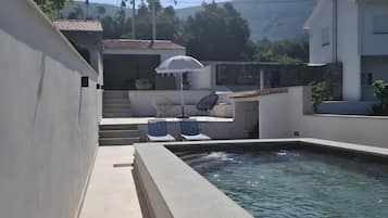 Une piscine bordée de pierres, deux chaises longues et un escalier menant à un espace abrité sous un parasol blanc.