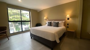 A modern bedroom with a large bed, two bedside lamps, a view through a large window, and a small seating area.