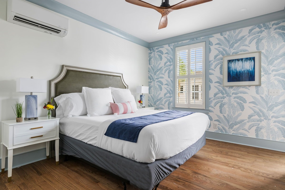 Quarto com cama, mesas de cabeceira, ventilador de teto, janela com persianas e parede com padrão floral.