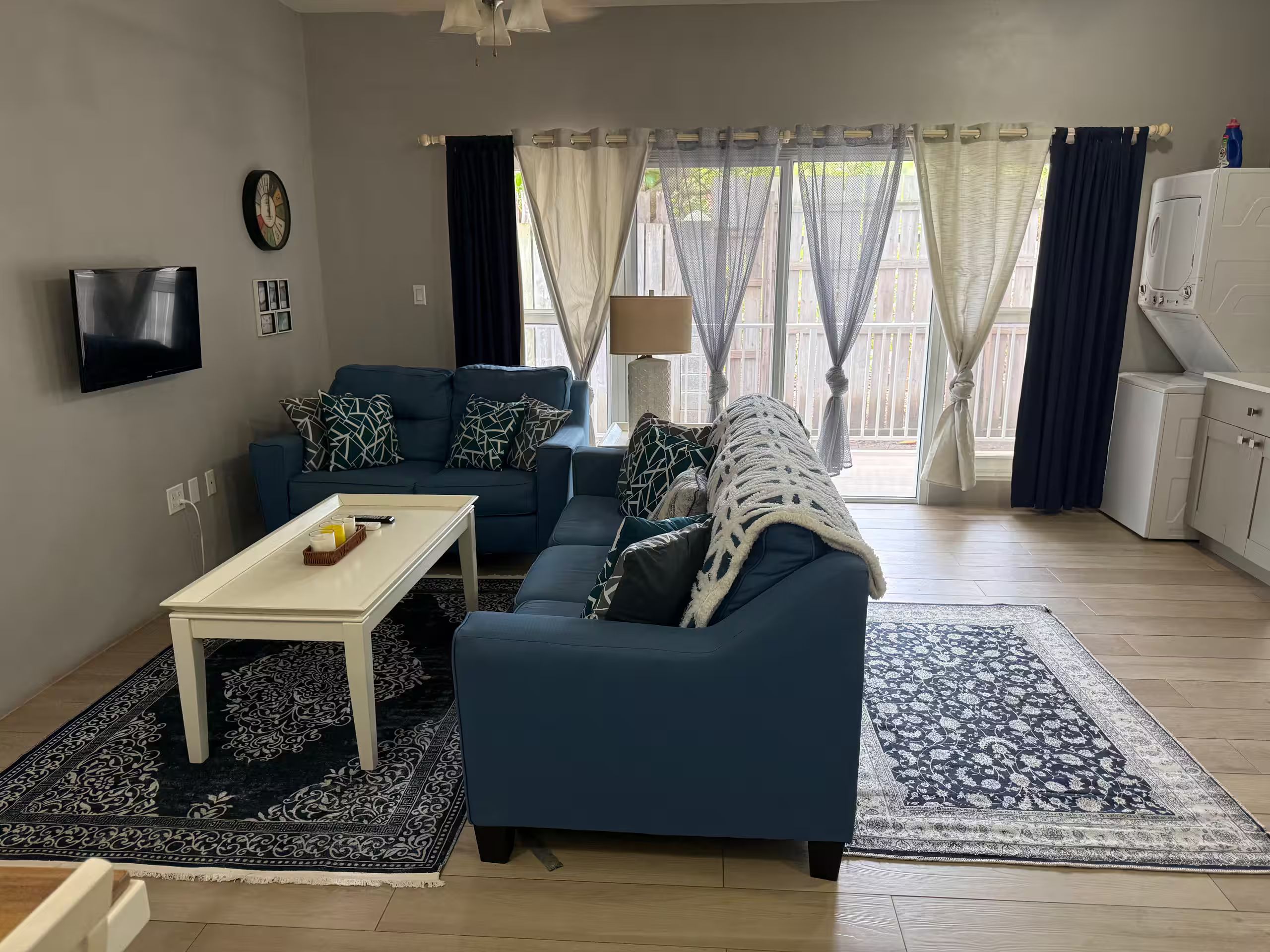 Een woonkamer met een blauwe bank, een witte salontafel, een gekleurd vloerkleed, een televisie en een raam met gordijnen.