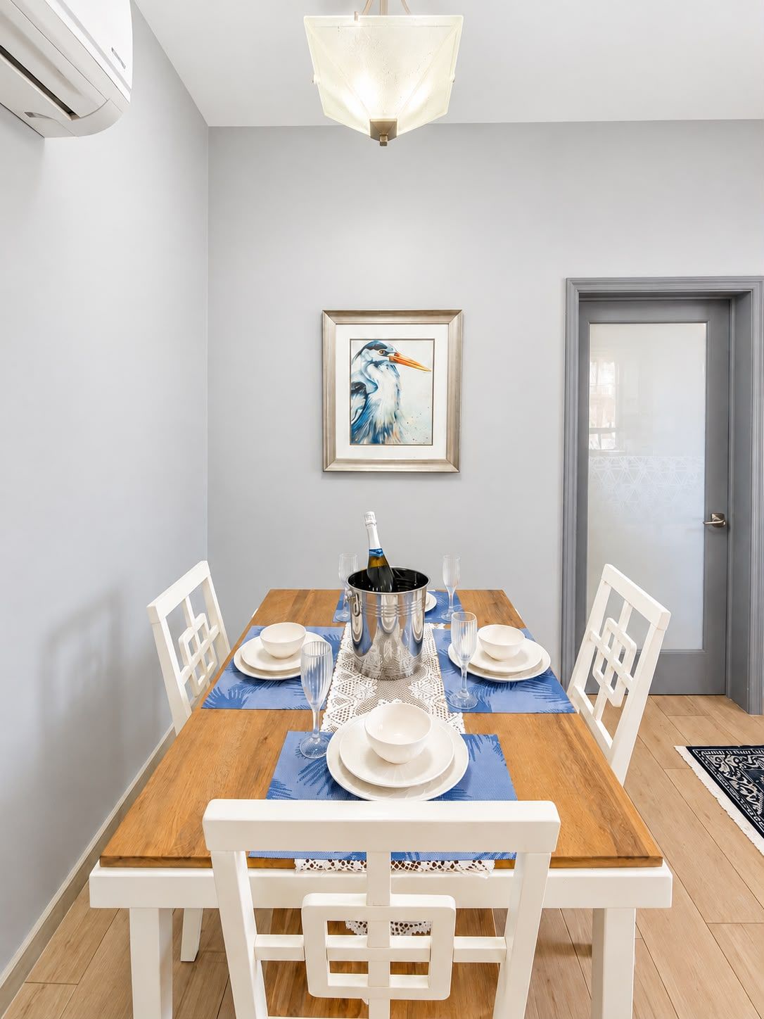 A dining room with a wooden table set for six, white chairs, a framed bird painting on the wall, and a door in the background.