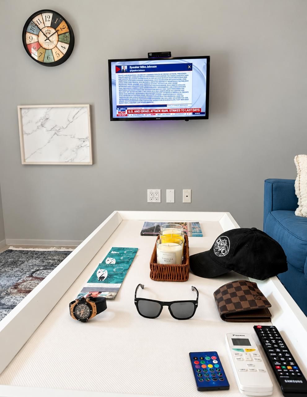 A room with a television displaying a news channel, a clock, a framed picture, a tray with personal items, and a blue couch.