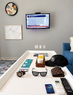 Une pièce avec un téléviseur diffusant une chaîne d’information, une horloge, un tableau encadré, un plateau contenant des effets personnels et un canapé bleu.