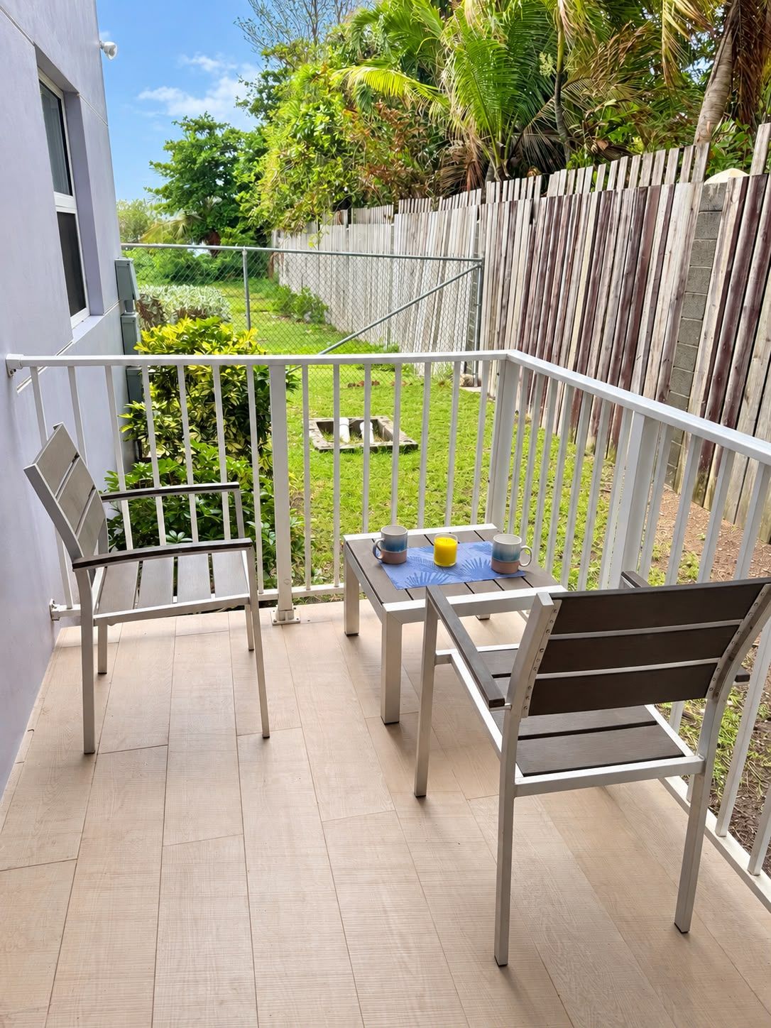 A balcony with a table and two chairs, a wooden fence, and a grassy yard.