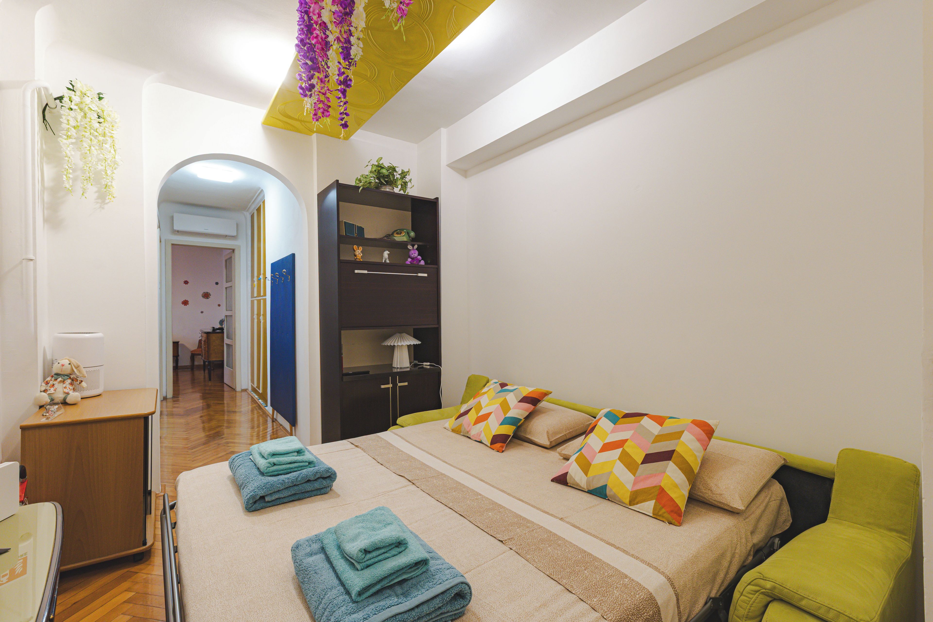 A bedroom with a bed, pillows, a nightstand, a dresser, a mirror, and a hanging plant.
