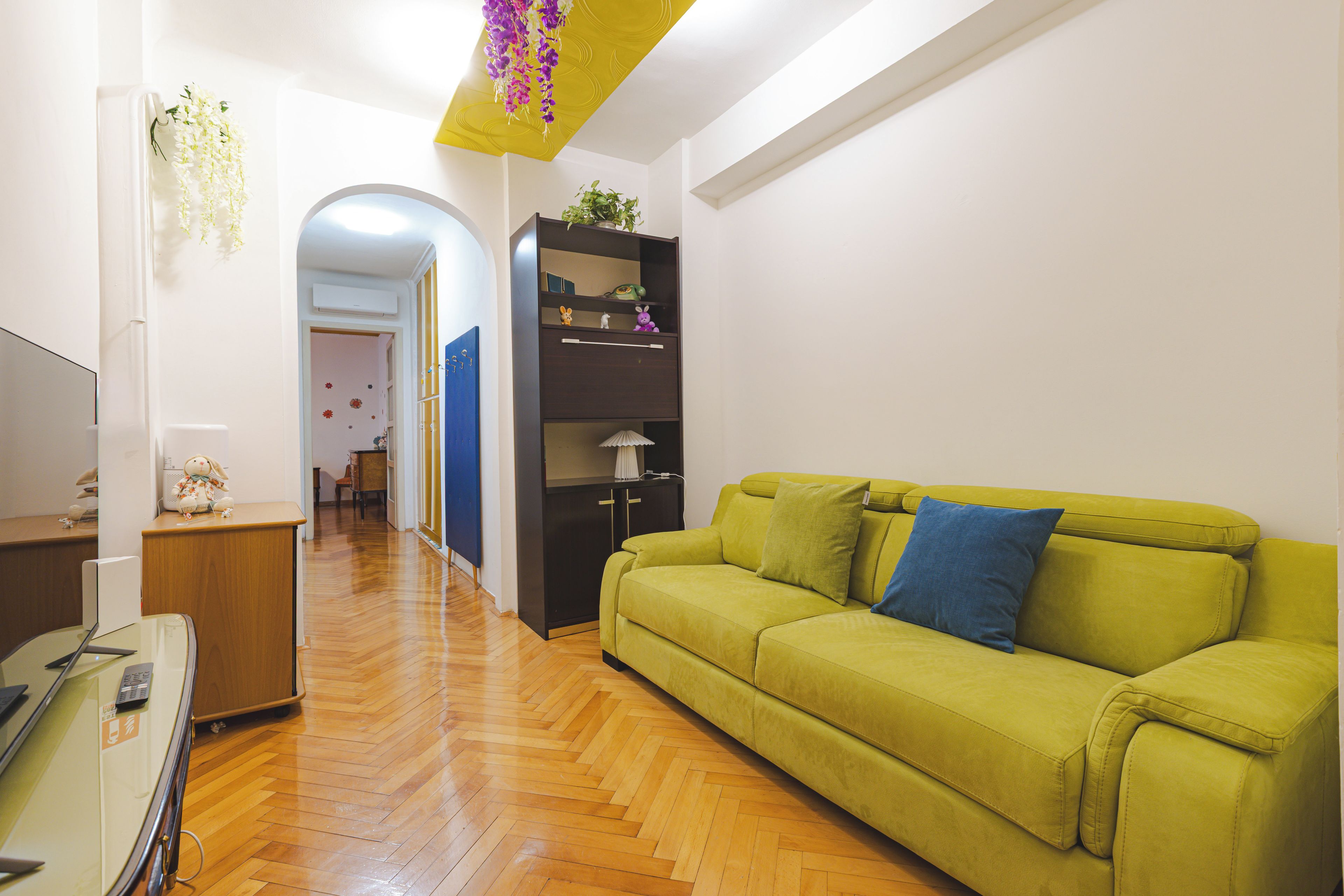 A room with a green sofa, wooden flooring, a desk with a computer, and a bookshelf with decorative items.