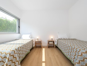 A bedroom with two beds, each with a patterned bedspread, wooden nightstands with lamps, and a window providing a view of greenery outside. - Hibiscus Bliss - Luxury villa V4 with private pool (Tavira)