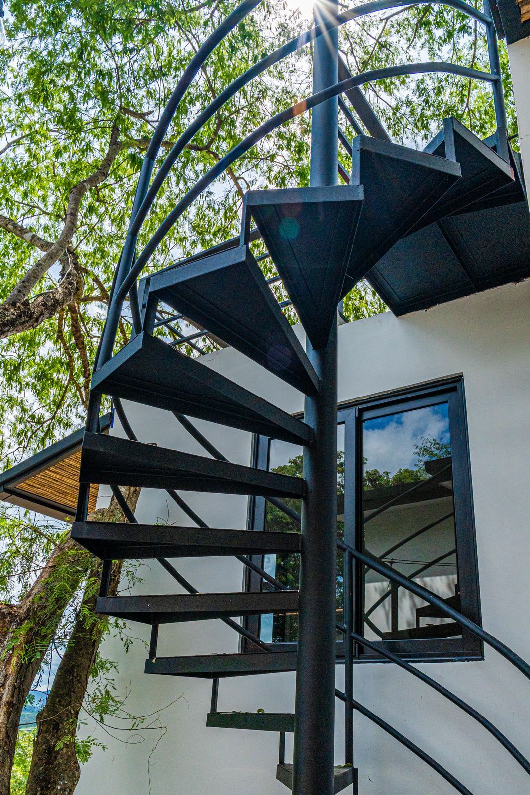 A spiral staircase with a metal frame and glass panels, leading to a building with a window.