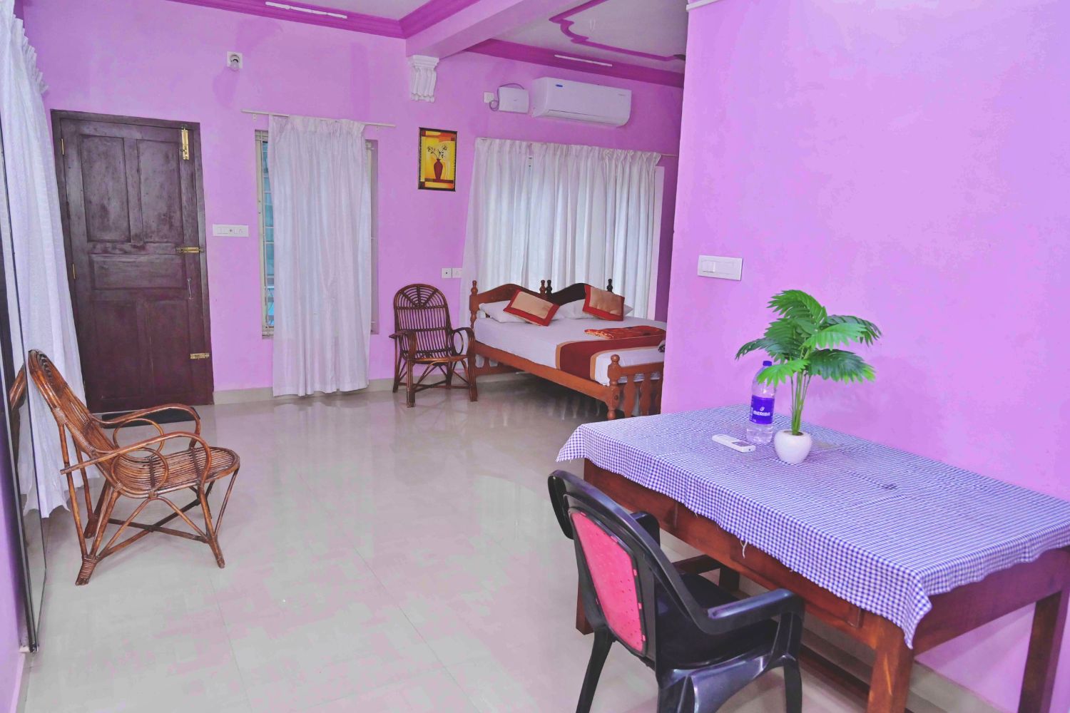 Una habitación con una cama, una mesa de comedor, dos sillas, una planta en maceta y un ventanal con cortinas.