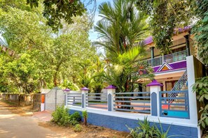 Una propiedad residencial con entrada protegida por un portón, una escalera que conduce a un balcón y abundante vegetación alrededor de la casa.