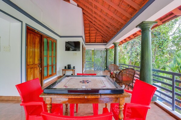 A room with a wooden table and red chairs, a television on the wall, and a balcony with a view of greenery. - Harbour villa, Vellayani Lake (Thiruvananthapuram)