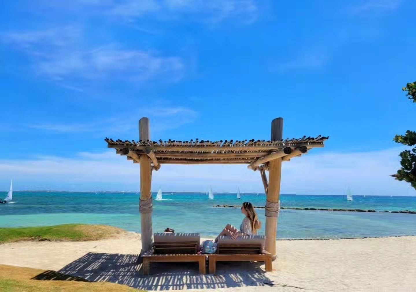 Ein Kapsel über dem Strand mit zwei Liegestühlen, einem Holzboden und Blick auf das Meer mit Segelbooten.