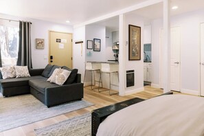 Uma sala de estar moderna com sofá seccional, área de jantar com banquetas, cozinha com armários brancos e uma cama grande.