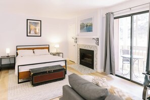 A bedroom with a bed, bedside tables, a fireplace, a balcony with outdoor furniture, and a large window. - Remodeled Mammoth Village Studio W/ Self Check In (Mammoth Lakes)