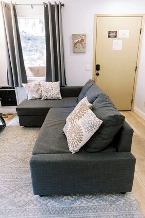 A modern living room with a grey sofa set, patterned cushions, a view through a window with curtains, and a door with a sign. - Remodeled Mammoth Village Studio W/ Self Check In (Mammoth Lakes)