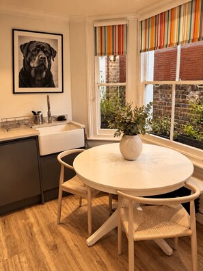 Uma cozinha com pia, um quadro com a imagem de um cachorro, uma mesa redonda com cadeiras e uma janela com cortinas listradas.