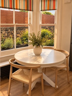A round table with a white surface and a vase with green plants in the center, surrounded by four wooden chairs with woven seats. - DR15 - Stylish 2-Bed with Patio near Queen's Park (Greater London)
