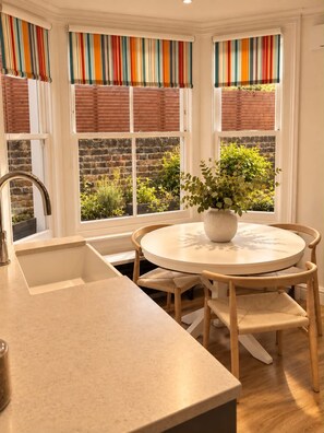 A kitchen with a round table, wooden chairs, a potted plant, and a window with a brick wall view. - DR15 - Stylish 2-Bed with Patio near Queen's Park (Greater London)