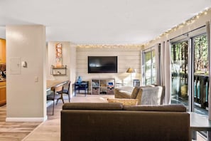 A modern living room with a flat-screen TV, a sofa, a dining table, and a view of a forest through large windows. - Pet Friendly Mammoth Studio w/ self check in (Mammoth Lakes)