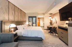 Ein modernes Hotelzimmer mit Bett, Nachttisch, Lampe, Sessel, Schreibtisch und Fernseher.