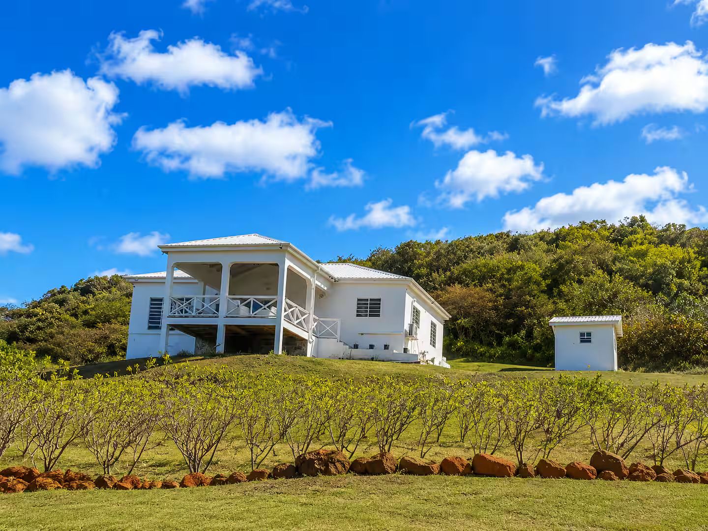 A white building with a balcony, surrounded by a vineyard and a hilly landscape.