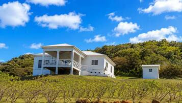 A white building with a balcony, surrounded by a vineyard and a hilly landscape.