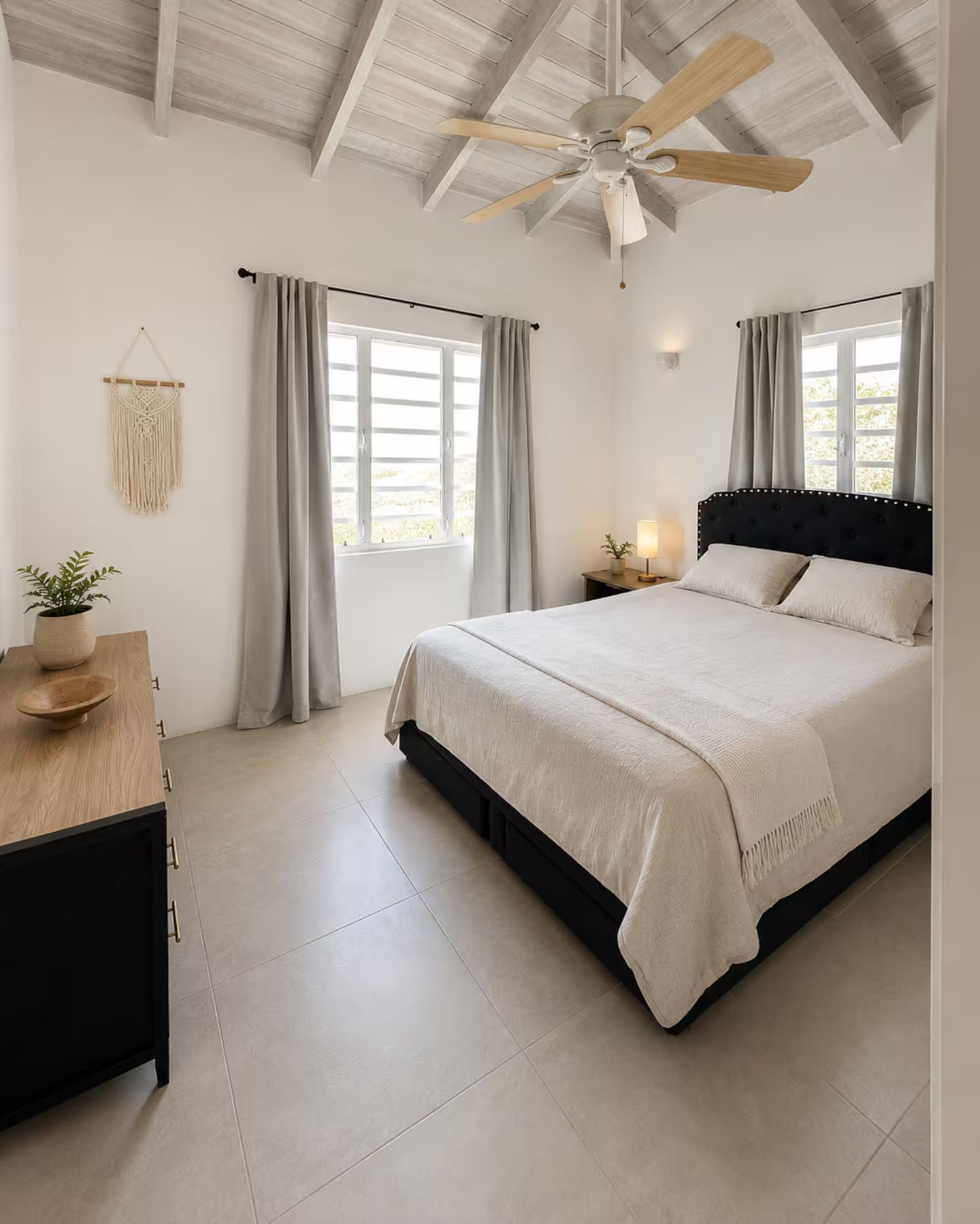 A bedroom with a bed, a ceiling fan, a nightstand, a potted plant, and a macrame wall hanging.
