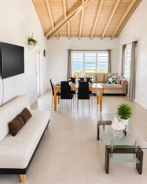 Un salon moderne comprenant un coin repas, un canapé, une table basse en verre et un téléviseur fixé au mur.