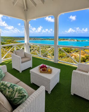 Un espace extérieur abrité, aménagé avec des meubles en osier et une table garnie de plats, offrant une vue sur un paysage côtier.