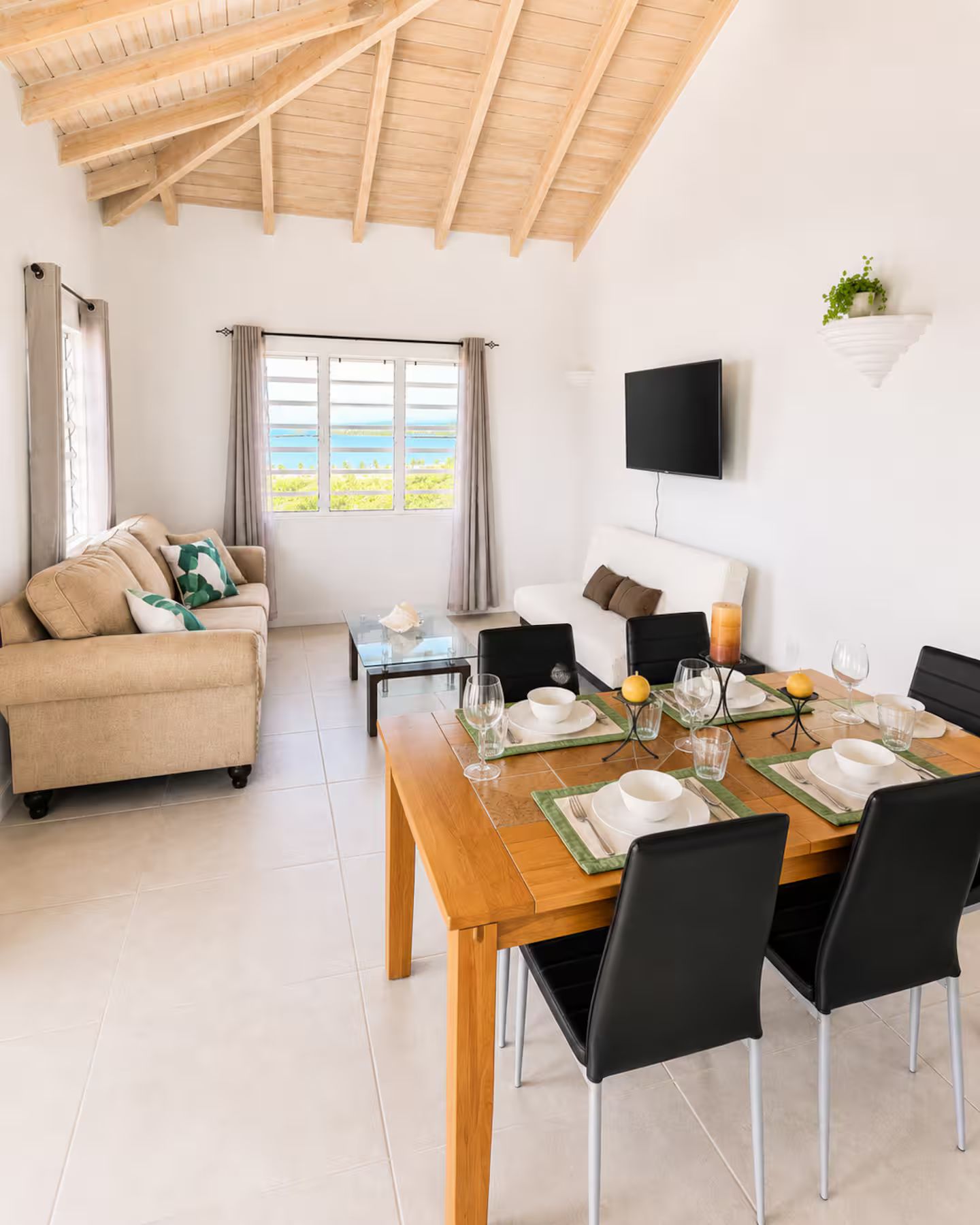 A modern living room with a wooden ceiling, a sofa, a dining table set for four, and a TV mounted on the wall.