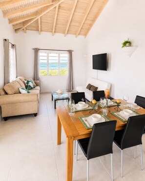 A modern living room with a wooden ceiling, a sofa, a dining table set for four, and a TV mounted on the wall.