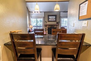 A dining area with a black marble table, wooden chairs, pendant lights, a fireplace, and a view of the outdoors. - Mammoth Home-Away-From-Home (Mammoth Lakes)