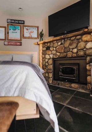 A bedroom with a stone fireplace, a bed with a grey blanket, and a flat-screen TV mounted above the fireplace. - Mammoth’s Modern Studio w/ Self Check in (Mammoth Lakes)