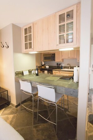 A kitchen with wooden cabinets, a microwave, a stove, and a bar area with stools. - Mammoth’s Modern Studio w/ Self Check in (Mammoth Lakes)