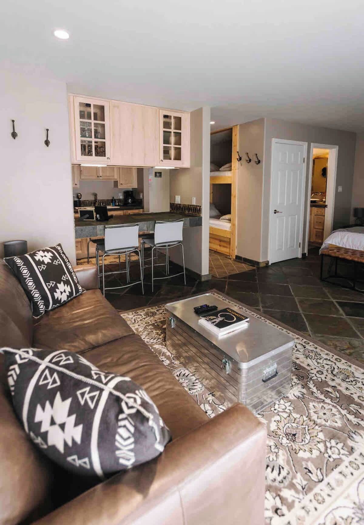 A compact living space with a sofa, coffee table, and patterned rug. A kitchenette is visible in the background.
