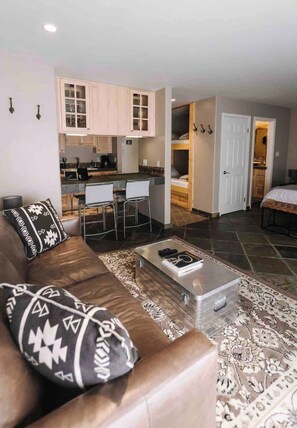 A compact living space with a sofa, coffee table, and patterned rug. A kitchenette is visible in the background. - Mammoth’s Modern Studio w/ Self Check in (Mammoth Lakes)