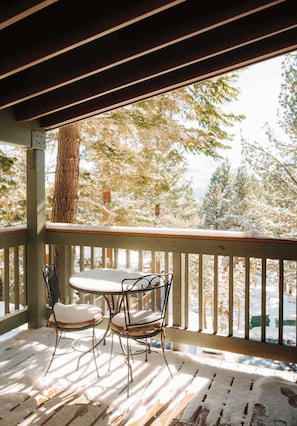 A covered outdoor seating area with a table and two chairs, overlooking a snowy landscape with trees. - Charming Village Studio Loft (Mammoth Lakes)