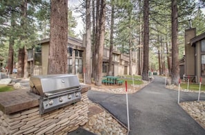 Een lodge met een stenen barbecue, een picknicktafel en een verhard pad, omringd door hoge bomen.