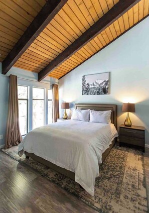 A bedroom with a large bed, wooden ceiling beams, a mountain landscape painting, and two bedside lamps. - Luxury Mammoth 2BR 2BA Townhome (Mammoth Lakes)