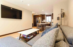 A modern living room with a flat-screen TV, a sofa, a coffee table with books, and a kitchen area in the background. - Mammoth Mountain Townhome w/ Garage EV Charger (Mammoth Lakes)