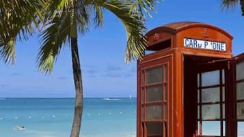 A red telephone booth on a beach with clear blue water and a palm tree.