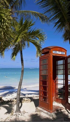 En rød telefonboks på en strand med klart, blåt vand og en palme.
