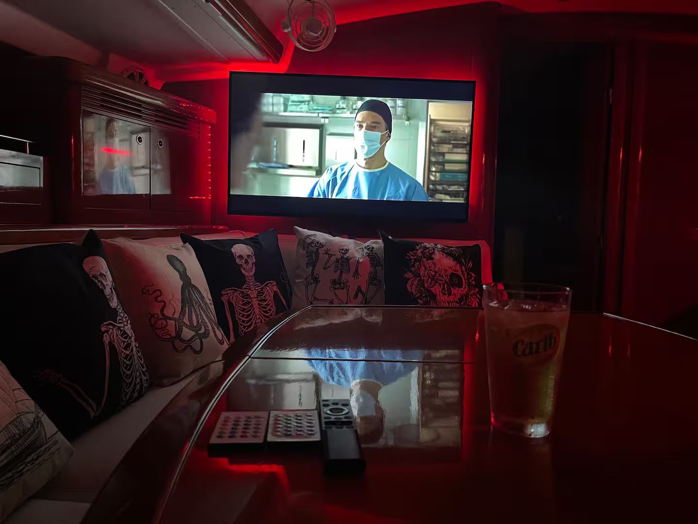 Ein Zimmer mit Fernseher an der Wand, einer Couch mit Skelett-Kissen, einer Fernbedienung und einem Bierglas auf einem Tisch.