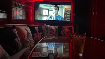 Ein Zimmer mit Fernseher an der Wand, einer Couch mit Skelett-Kissen, einer Fernbedienung und einem Bierglas auf einem Tisch.