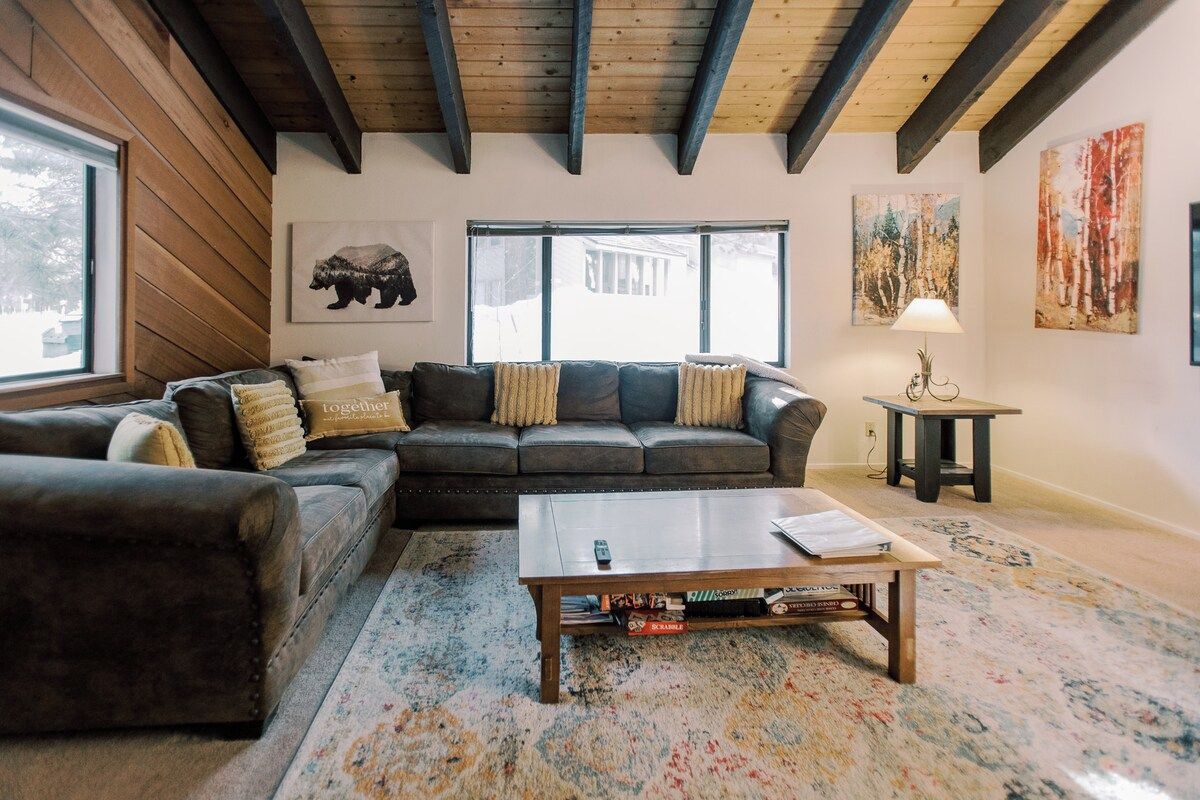 A living room with a sectional sofa, a coffee table, and a rug. There are large windows and artwork on the walls.