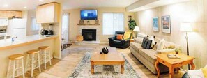 A living room with a sofa, a coffee table, and a TV. There is a kitchen area with stools and a fireplace. - Mammoth Horizon 4 1BD 1BA Family Getaway (Mammoth Lakes)