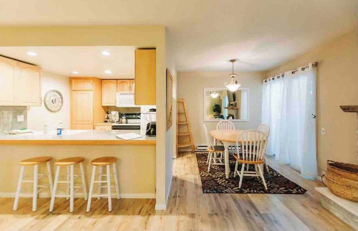 A kitchen with wooden cabinets, a central island with stools, a dining area with a table and chairs, and a staircase leading to another level.