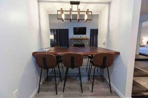 Een eetruimte met een lange tafel en vier stoelen, een open haard en een aan de muur bevestigde televisie.